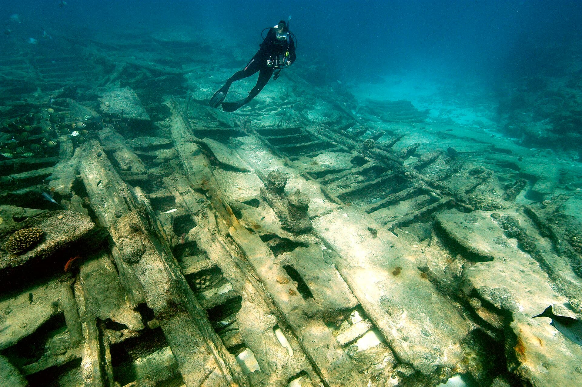 A diver with a camera glides over the broken iron frames of a wreck on a sandy bottom